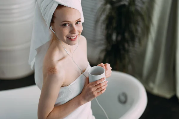 Счастливая женщина в ванной слушает музыку и пьет чай сидя на ванне — стоковое фото