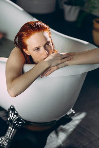 Mujer joven pelirroja acostada en la bañera, tocándose suavemente la cara con las manos — Foto de Stock