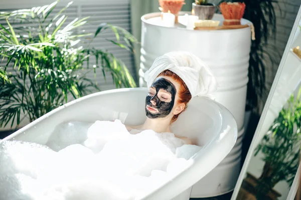 Donna con un asciugamano in testa sdraiata nella vasca da bagno con una maschera di argilla sul viso — Foto Stock