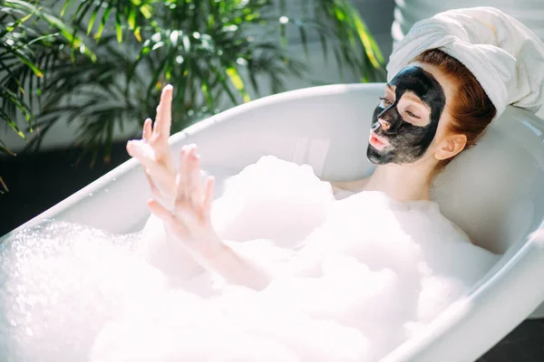 Femme avec une serviette sur la tête couchée dans la baignoire avec un masque d'argile sur le visage — Photo