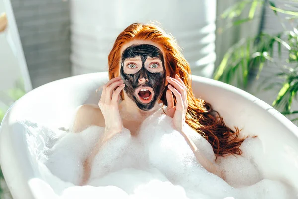Mujer con el pelo largo y rojo acostada en la bañera con una máscara de barro en la cara —  Fotos de Stock