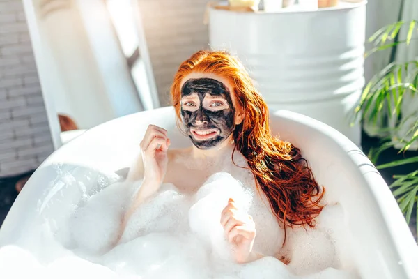 Vrouw met lang rood haar liggend in bad met een klei masker op haar gezicht — Stockfoto