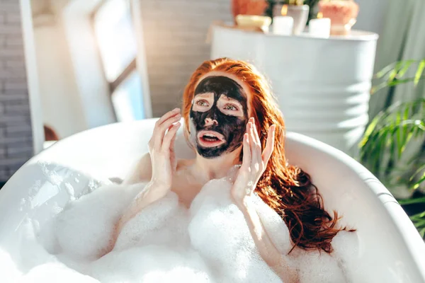 Donna con lunghi capelli rossi sdraiata nella vasca da bagno con una maschera di argilla sul viso — Foto Stock