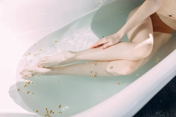 Foto recortada de piernas delgadas femeninas en bañera con agua limpia y pequeñas flores — Foto de Stock