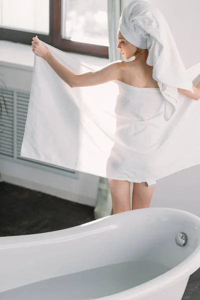 Modelo femenino parado entre ventana y bañera, usando toalla para ocultar su desnudez — Foto de Stock