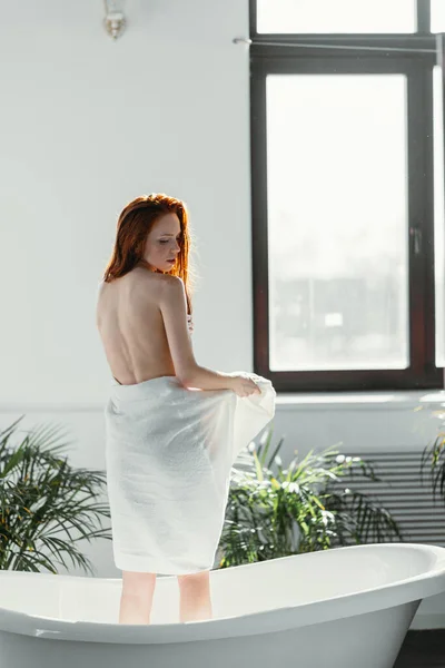 Kvinnlig modell stående mellan fönster och badkar, med handduk för att dölja sin nakenhet — Stockfoto