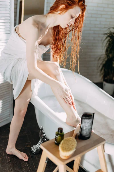 Young woman applying coffee scrub onto her legs in bathroom