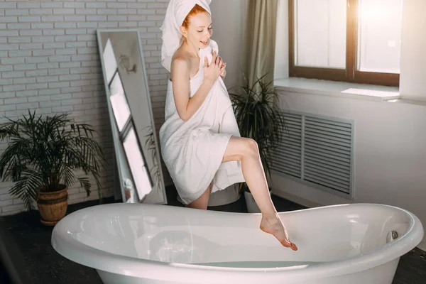 욕조 가장자리에 앉아 목욕을 하고 손으로 물을 시험하는 미소짓는 여자 — 스톡 사진