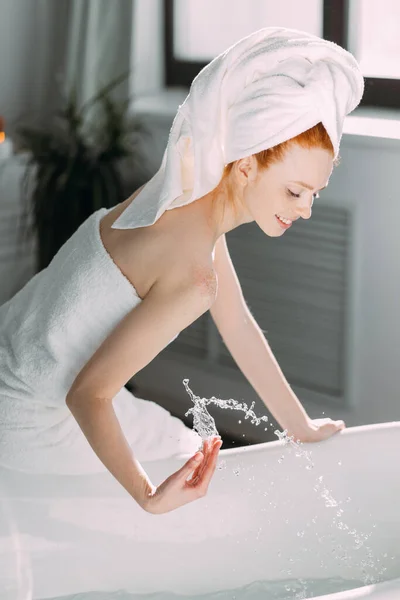 Mujer sonriente sentada en el borde de la bañera corriendo el baño, probando el agua con la mano —  Fotos de Stock