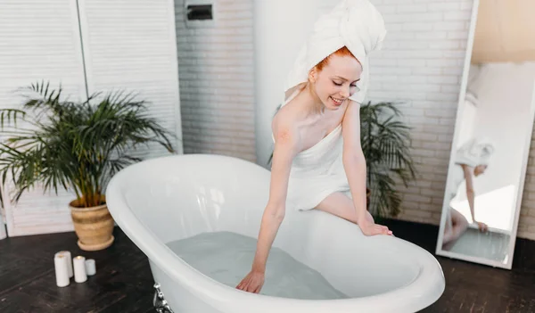 Leende kvinna som sitter på badkarskanten och badar, testar vatten med handen — Stockfoto