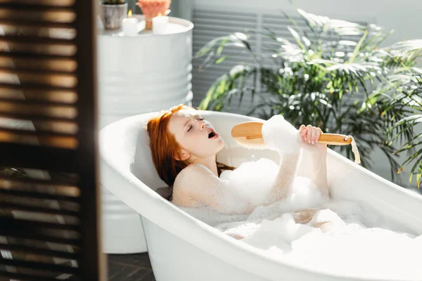 Joyful vrouw zingen met behulp van borstel als microfoon ontspannen in bad in de badkamer — Stockfoto