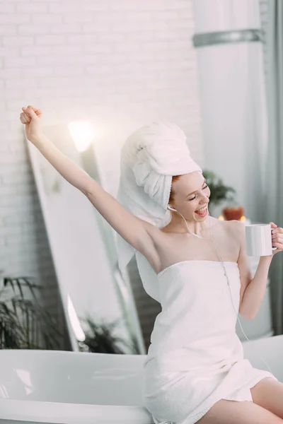 Glad kvinna i badrummet lyssnar på musik och dricker te sittande på badkar — Stockfoto