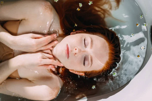 Κοκκινομάλλα νεαρή γυναίκα ξαπλωμένη στην μπανιέρα, ακουμπώντας απαλά το πρόσωπό της με τα χέρια — Φωτογραφία Αρχείου