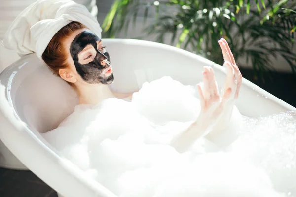 Donna con un asciugamano in testa sdraiata nella vasca da bagno con una maschera di argilla sul viso — Foto Stock