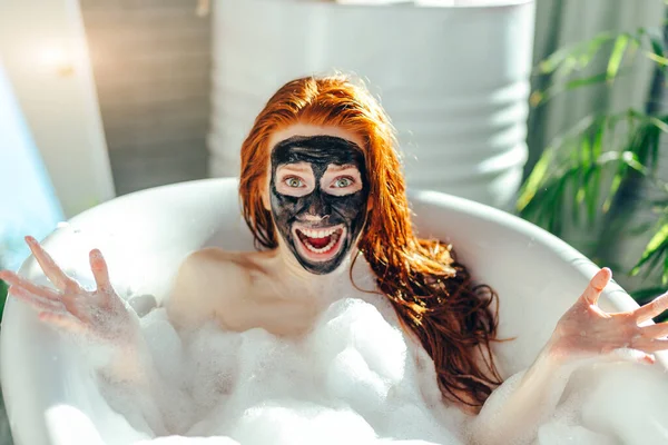 Vrouw met lang rood haar liggend in bad met een klei masker op haar gezicht — Stockfoto