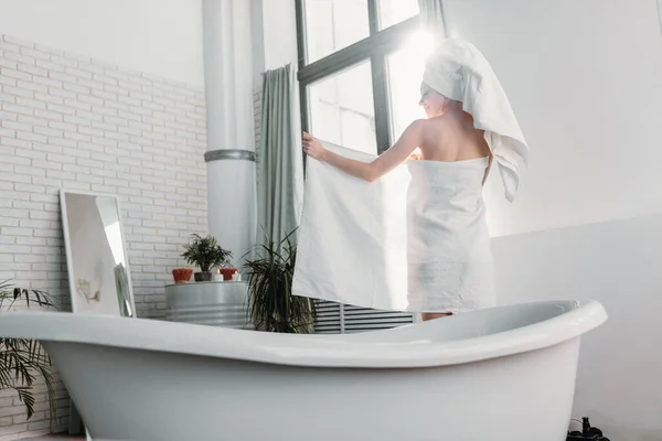 Mulher de cabelos vermelhos magro cobrindo seu corpo com toalha enquanto está de pé no banheiro — Fotografia de Stock