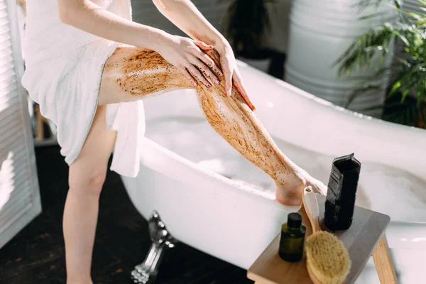 Mujer joven aplicando un exfoliante de café en sus piernas en el baño — Foto de Stock