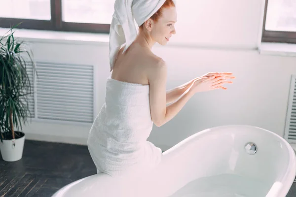 Mujer sonriente sentada en el borde de la bañera corriendo el baño, probando el agua con la mano — Foto de Stock