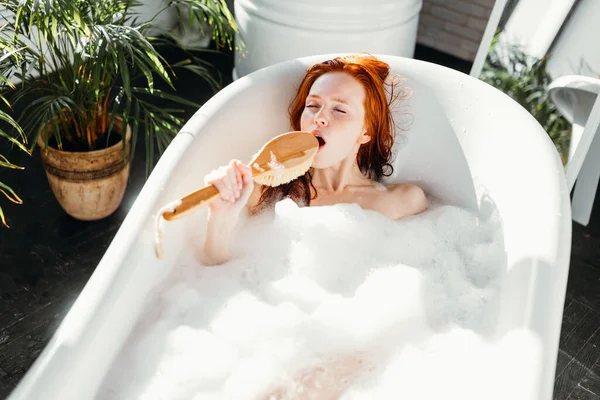 Радісна жінка співає, використовуючи щітку, як мікрофон, що розслабляється у ванній — стокове фото
