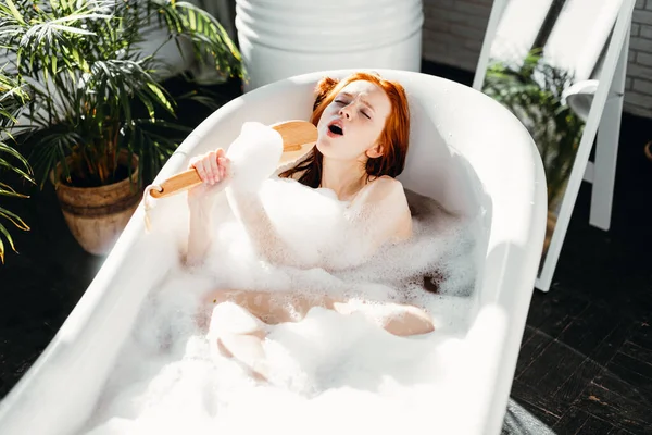 Mujer alegre cantando usando cepillo como micrófono relajante en baño en baño — Foto de Stock