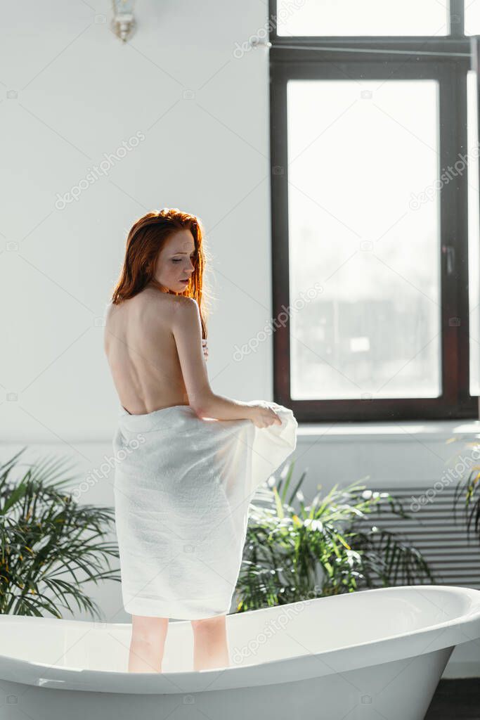 Female model standing between window and bathtub, using towel to hide her nudity