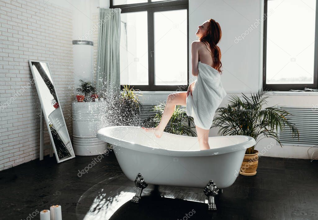 Red-haired slim woman covering her body with towel while standing in bathroom