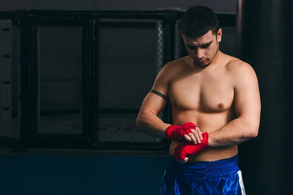 Boxeador masculino sin camisa en pantalones cortos azules envuelve sus nudillos con vendaje elástico —  Fotos de Stock