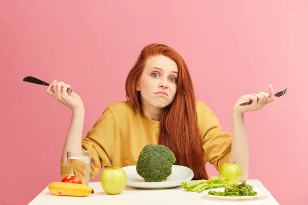 Kafası karışmış, belirsiz kızıl saçlı bir kadın masada oturuyor, elinde brokoli var. — Stok fotoğraf