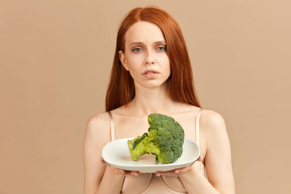 Anorexic woman in underwear holding plate with raw broccoli isolated in studio