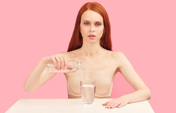 Anorexic déterminé femme se trouve dans les boissons sous-vêtements que de l'eau contre le mur rose. — Photo