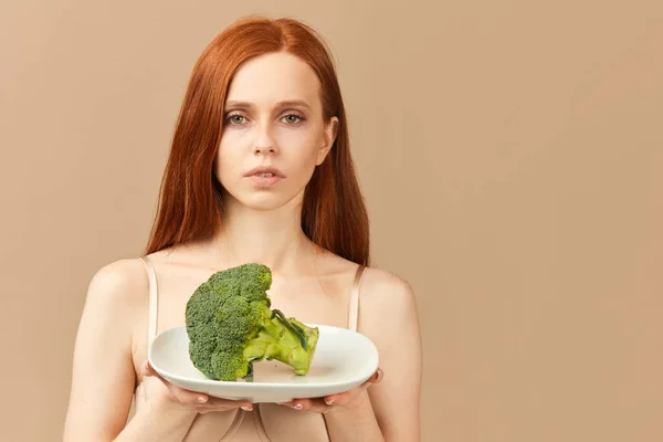 Anorexia vrouw in ondergoed holding plaat met ruwe broccoli geïsoleerd in studio — Stockfoto