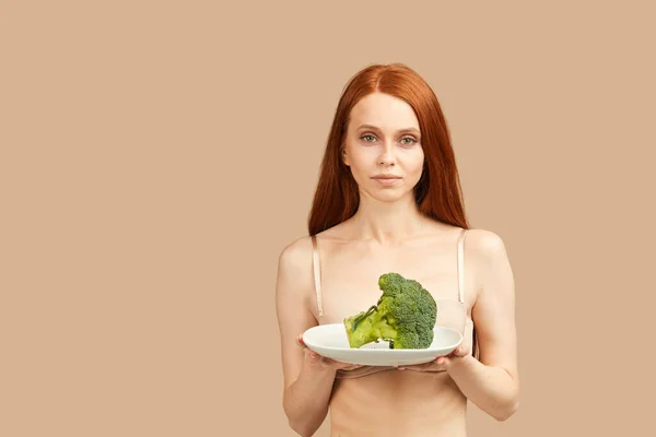 Anorektyczka w bieliźnie trzymająca płytkę z surowymi brokułami izolowana w studio — Zdjęcie stockowe