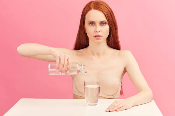 Anorexic determined woman sits in underwear drinks only water against pink wall.