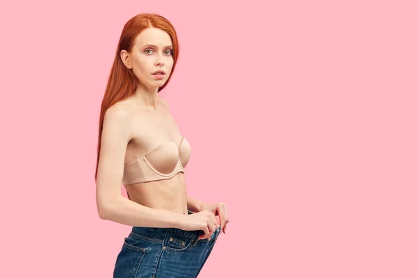 Ung smal anorektisk kvinna i överdimensionerade jeans visar hennes diet resultat på rosa — Stockfoto