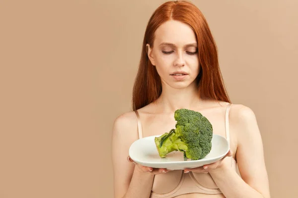 Anorexická žena ve spodním prádle drží talíř se syrovou brokolicí izolované ve studiu — Stock fotografie