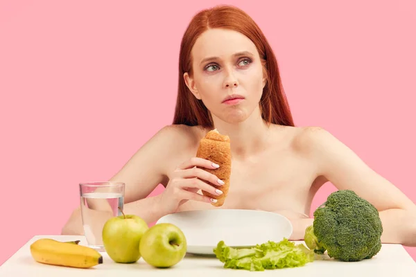 Nudí apatická žena jí croissant zírá do vesmíru ignoruje syrové ovoce — Stock fotografie