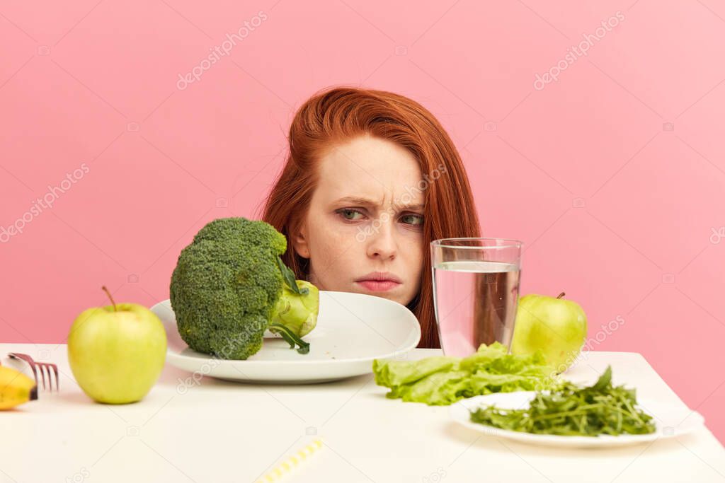 Frowning woman dislikes vegetables on table with disgusting grimace isolated