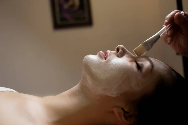 目を閉じて化粧品師と寝そべっている女性が顔のマスクをブラシで — ストック写真