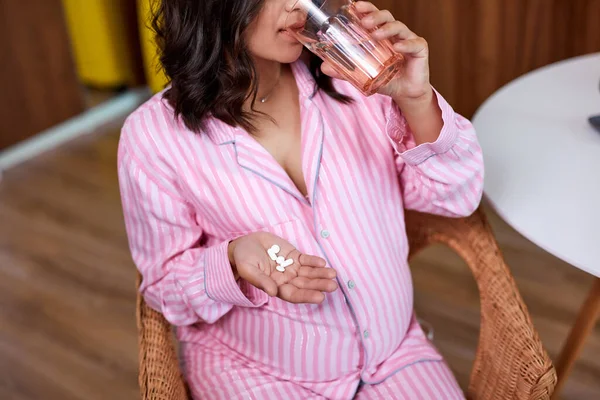 Mulher grávida atraente tomando um comprimido sentado em um sofá na sala de estar em casa — Fotografia de Stock