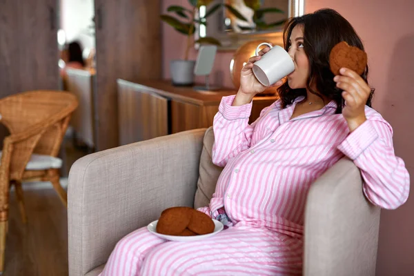 Mulher grávida está comendo biscoitos de chocolate e bebendo chá em casa — Fotografia de Stock