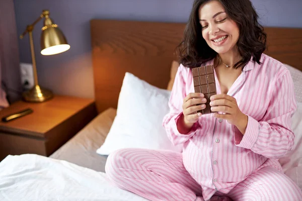 Pregnant woman enjoys eating chocolate in bed at home — Stock Photo, Image