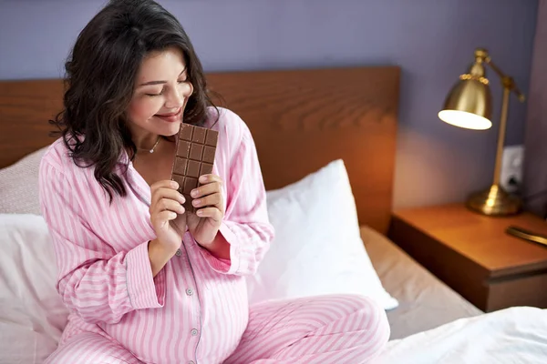 Smiling playful pregnant woman eating chocolate bar on bed — Stock Photo, Image