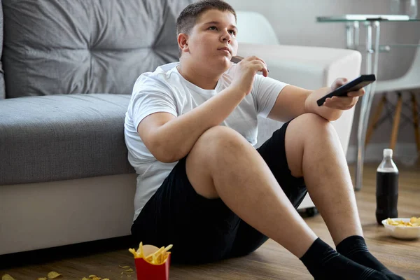 Otyły chłopiec jeść niezdrowe jedzenie podczas oglądania telewizji sam w domu — Zdjęcie stockowe