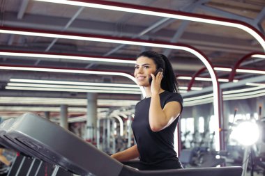 Kadın koşu bandında koşarken cep telefonuyla konuşurken gülümsüyor.