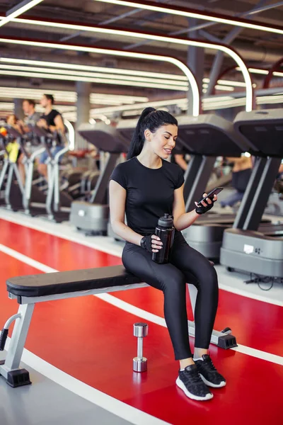 Fit γυναίκα με μπουκάλι χρησιμοποιώντας το έξυπνο τηλέφωνό της με ακουστικά στο γυμναστήριο — Φωτογραφία Αρχείου