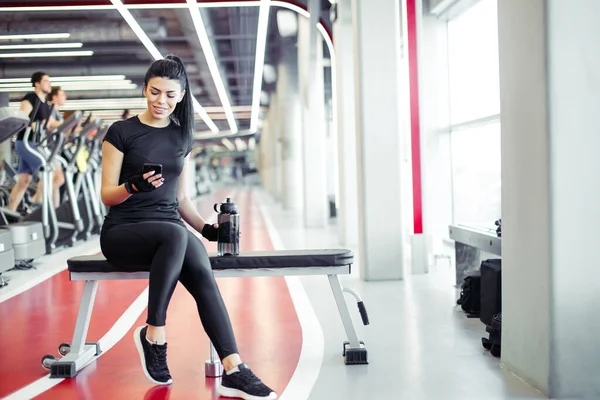 Ajuste mujer con botella usando su teléfono inteligente con auriculares en el gimnasio — Foto de Stock