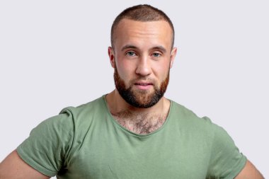 Beyaz arka planda sıradan giyinmiş yakışıklı sakallı adam kameraya bakıyor..