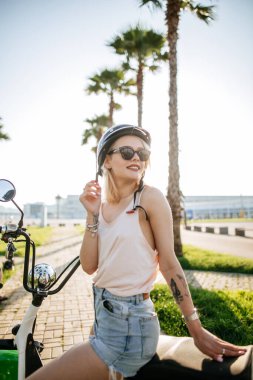 Kasklı ve koruyucu gözlüklü genç kız elektrikli bisikletin yanında oturuyor.