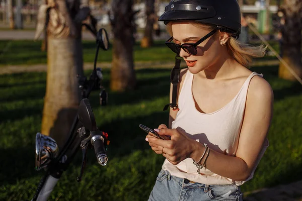 Attraktiv kvinna i cykelhjälm med elcykel i parken med telefon — Stockfoto