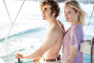 Kız arkadaşıyla bir yatın dümenini tutan ve uzaklara bakan bir adam.